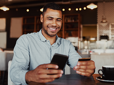 Young man sitting in coffee shop looking at cell phone and holding credit card in other hand