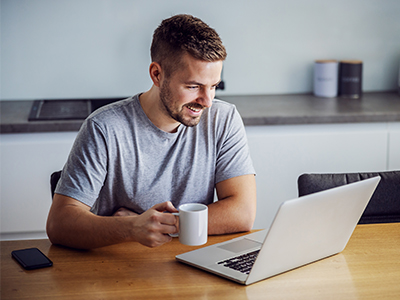 Man sitting at table holding coffee cup and looking at laptop screen