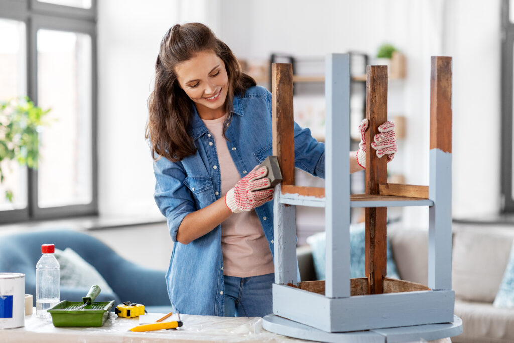 furniture renovation, diy and home improvement concept - happy smiling woman sanding old wooden table with sponge