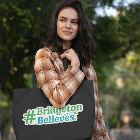 A woman in a plaid shirt holds a black tote bag with "#BridgetonBelieves" in green text. She stands outdoors with a calm expression, surrounded by trees.