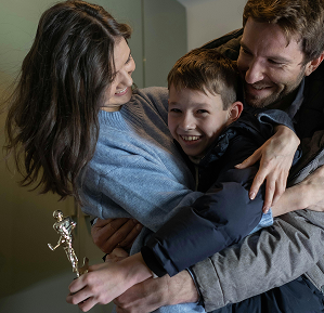 A joyful family hug with a smiling boy holding a trophy. The mother in a blue sweater and father in a gray jacket express pride and happiness.