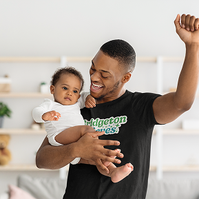 A smiling man holds a baby in one arm, raising the other in joy. He's wearing a black T-shirt with green text. The scene is warm and lively in a cozy room.