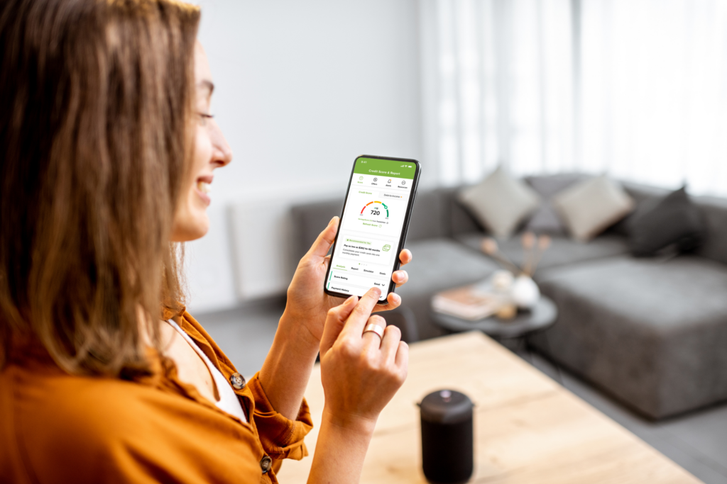 A smiling woman in an orange shirt uses a smartphone app showing a credit score. She's in a modern living room with a gray sofa in the background.
