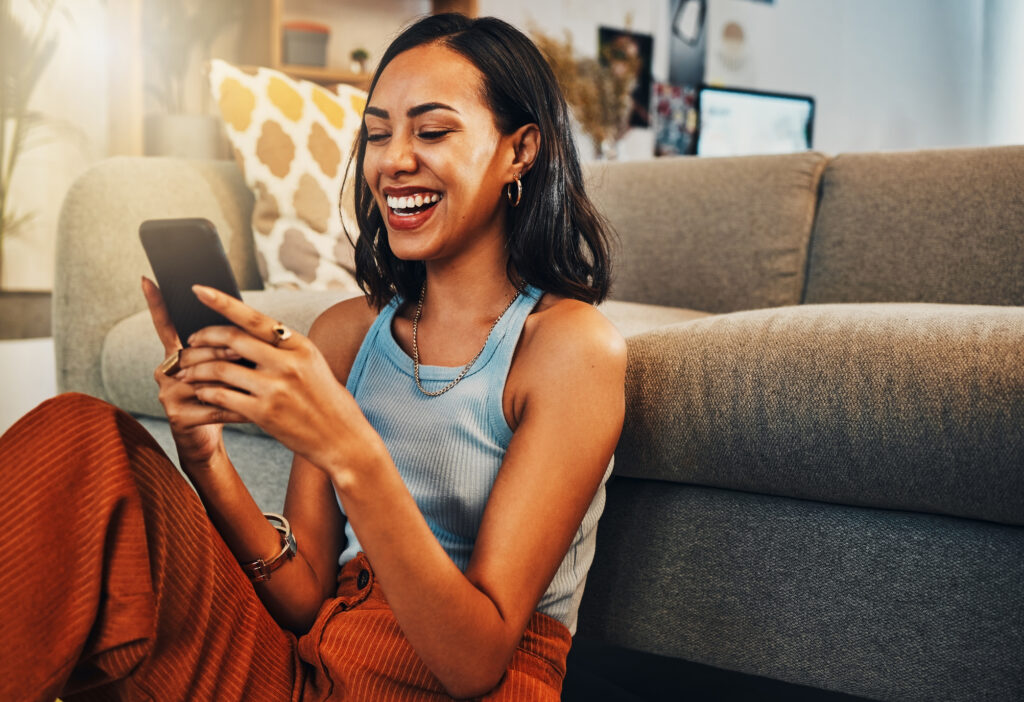 A woman joyfully laughs while looking at her smartphone, seated on a living room floor against a sofa. The setting is cozy and relaxed.